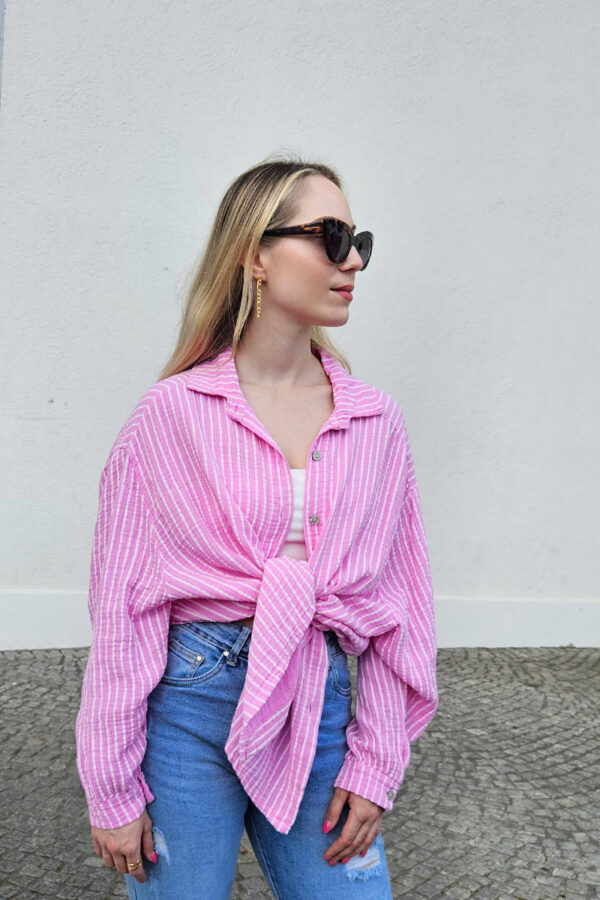 Gestreifte, lockere Bluse in rosa aus 100% Baumwolle, gefertigt aus weichem Musselinstoff. Die Bluse hat einen Oversize-Schnitt, der für einen lässigen Look sorgt. Sie besitzt einen V-Ausschnitt mit Umlegekragen und eine durchgehende Knopfleiste. Hinten ist eine querlaufende Falte eingearbeitet, die für zusätzliche Struktur sorgt. Die Bluse hat eine mittlere Länge und lange Ärmel mit 1-Knopf-Manschetten an den Ärmelenden.