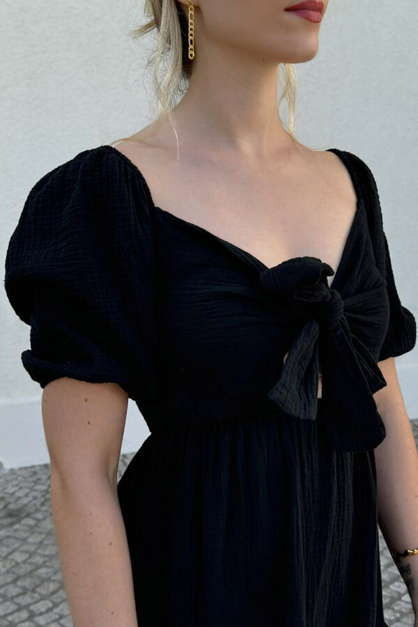 Ein verspieltes und bequemes Kleidchen in schwarz aus 100% Baumwolle, gefertigt aus leichtem Musselinstoff. Das Kleid hat einen Gummizug an der Taille und an den kurzen Ärmeln, was für eine bequeme Passform sorgt. Der Rücken ist gesmokt und das Oberteil ziert eine Schleife zum Binden, die sowohl vorne als auch hinten getragen werden kann. Der Saum ist mit einem verspielten Volant versehen, der dem Kleid eine leichte und luftige Note verleiht. Dieses Kleid ist perfekt für den Sommer geeignet.