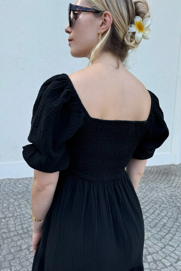 Ein verspieltes und bequemes Kleidchen in schwarz aus 100% Baumwolle, gefertigt aus leichtem Musselinstoff. Das Kleid hat einen Gummizug an der Taille und an den kurzen Ärmeln, was für eine bequeme Passform sorgt. Der Rücken ist gesmokt und das Oberteil ziert eine Schleife zum Binden, die sowohl vorne als auch hinten getragen werden kann. Der Saum ist mit einem verspielten Volant versehen, der dem Kleid eine leichte und luftige Note verleiht. Dieses Kleid ist perfekt für den Sommer geeignet.