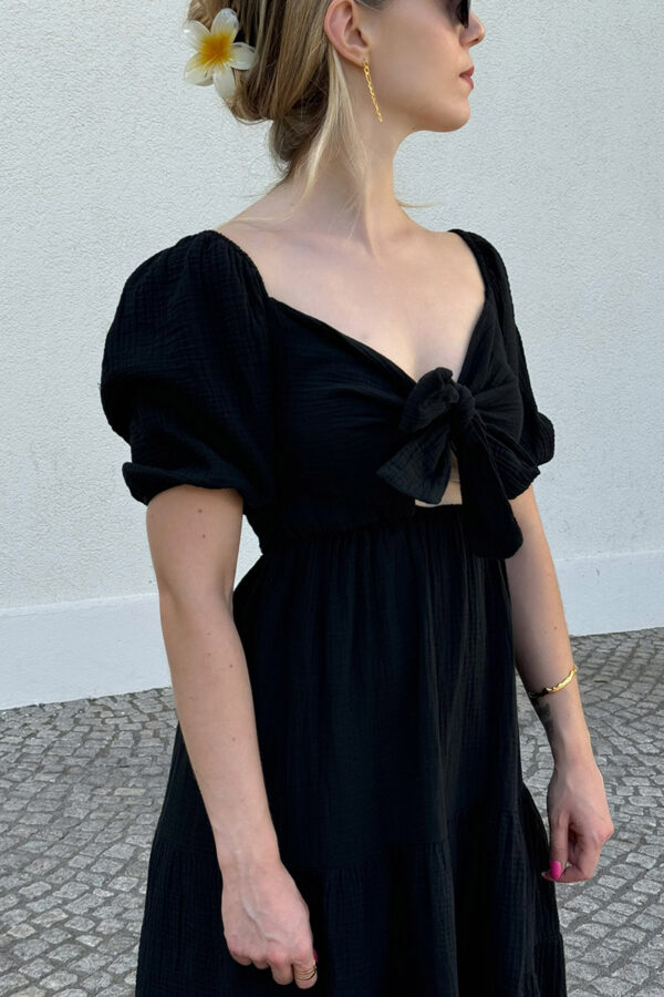 Ein verspieltes und bequemes Kleidchen in schwarz aus 100% Baumwolle, gefertigt aus leichtem Musselinstoff. Das Kleid hat einen Gummizug an der Taille und an den kurzen Ärmeln, was für eine bequeme Passform sorgt. Der Rücken ist gesmokt und das Oberteil ziert eine Schleife zum Binden, die sowohl vorne als auch hinten getragen werden kann. Der Saum ist mit einem verspielten Volant versehen, der dem Kleid eine leichte und luftige Note verleiht. Dieses Kleid ist perfekt für den Sommer geeignet.
