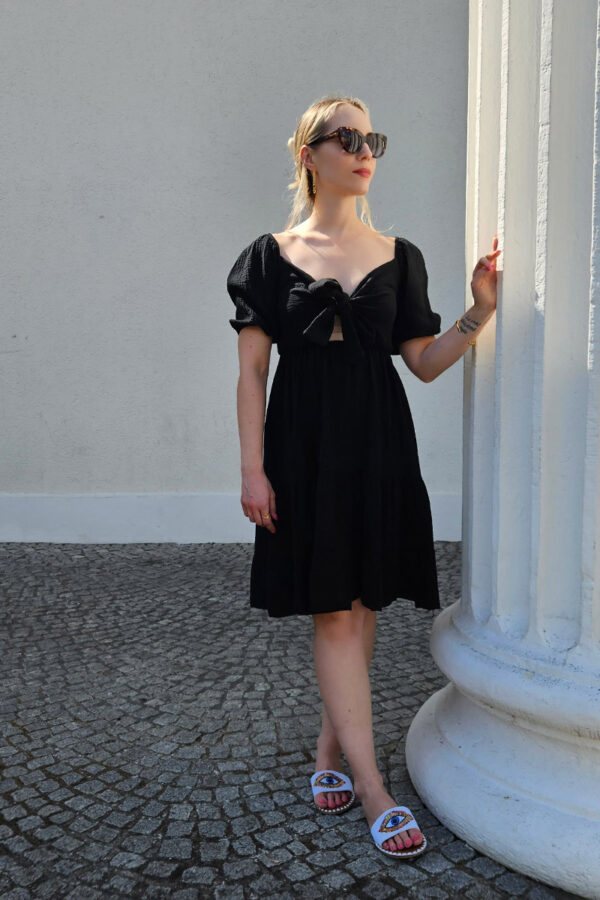 Ein verspieltes und bequemes Kleidchen in schwarz aus 100% Baumwolle, gefertigt aus leichtem Musselinstoff. Das Kleid hat einen Gummizug an der Taille und an den kurzen Ärmeln, was für eine bequeme Passform sorgt. Der Rücken ist gesmokt und das Oberteil ziert eine Schleife zum Binden, die sowohl vorne als auch hinten getragen werden kann. Der Saum ist mit einem verspielten Volant versehen, der dem Kleid eine leichte und luftige Note verleiht. Dieses Kleid ist perfekt für den Sommer geeignet.