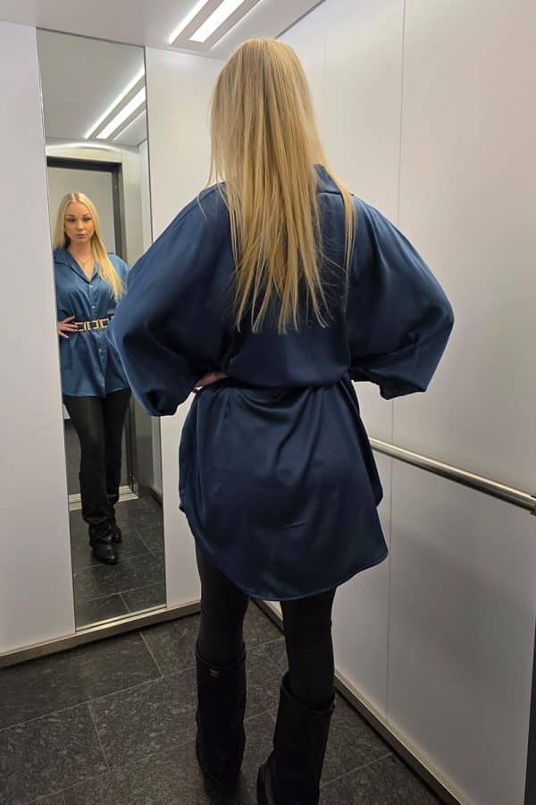 Dunkelblaue Oversize Damenbluse aus weich fließendem, leicht glänzendem Stoff. Die Bluse hat lange Fledermausärmel, eine klassische Knopfleiste vorne und einen dezenten Stehkragen. Der verlängerte, abgerundete Saum am Rücken sorgt für einen modernen, eleganten Look. Ideal für Büro, Alltag oder festliche Anlässe, mit lockerer Passform für maximalen Tragekomfort.