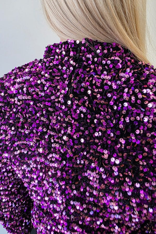 Hochwertiges, lila und pink schimmerndes Glitter- und Pailletten-Top mit langen Ärmeln und Schulterpolstern. Das Oberteil besitzt einen hohen Rundhalsausschnitt, einen figurbetonten Schnitt und einen dezenten Reißverschluss am Rücken. Die dicht gesetzten Pailletten reflektieren das Licht intensiv und verleihen dem Top einen starken Disco- und Festivallook.