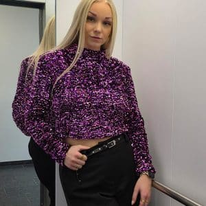 Hochwertiges, lila und pink schimmerndes Glitter- und Pailletten-Top mit langen Ärmeln und Schulterpolstern. Das Oberteil besitzt einen hohen Rundhalsausschnitt, einen figurbetonten Schnitt und einen dezenten Reißverschluss am Rücken. Die dicht gesetzten Pailletten reflektieren das Licht intensiv und verleihen dem Top einen starken Disco- und Festivallook.