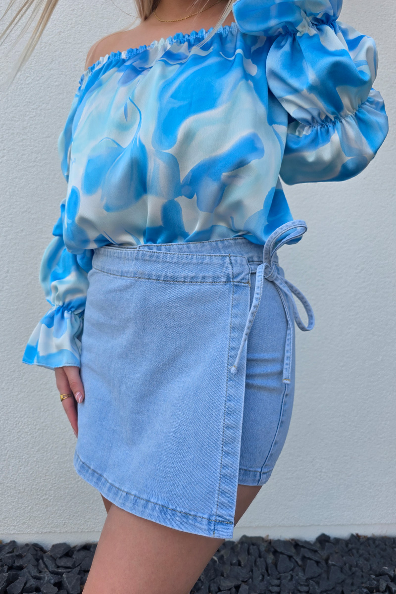 Stilvoller Jeans Skort im Wickel-Look aus hellem Denim-Blau mit seitlicher Schleife zum Binden, integriertem Reißverschluss und Metallknopf. Der Skort verfügt über praktische Hosentaschen hinten und eine kurze Innenhose für maximalen Komfort und volle Bewegungsfreiheit – perfekt für Citytrips, Festivals oder entspannte Tage am Strand.