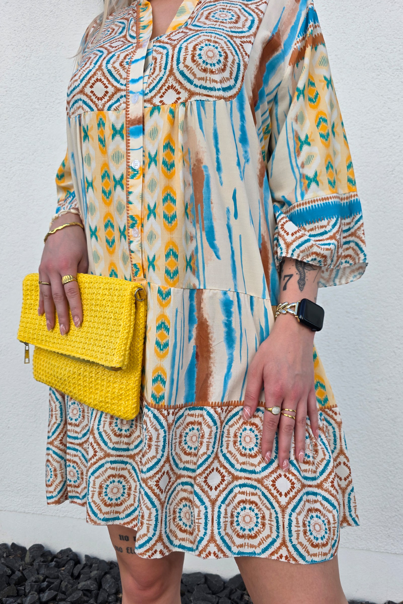 Dieses leichte Boho Kleid mit Knopfleiste ist aus luftigem, fließendem Stoff gefertigt und mit einem auffälligen Ethno-Muster in Blau, Braun und Gelb verziert. Der kurze, lockere Schnitt in knieumspielender Länge sorgt für Bewegungsfreiheit, während der V-Ausschnitt und die 3/4-Ärmel mit leichtem Puff-Effekt dem Kleid eine feminine und sommerliche Note verleihen.